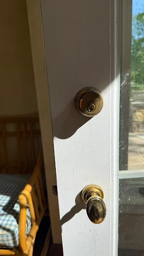 Deadbolt Installation by Local Locksmith Pros LLC, featuring a close-up of a brass deadbolt and matching doorknob on a white exterior door, illuminated by sunlight.